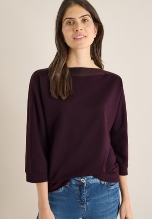 Burgundy long-sleeve top with a boat neckline, soft texture, and rolled cuffs, paired with light blue jeans featuring a button and pocket.