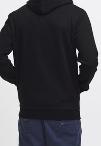 Felpa nera con cappuccio e tessuto morbido, dotata di tasca a marsupio e polsini a costine. Indossata con pantaloni blu navy.