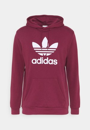 Personne portant un sweat à capuche Adidas bordeaux, un jean bleu clair et des baskets blanches, debout devant un fond clair uni.