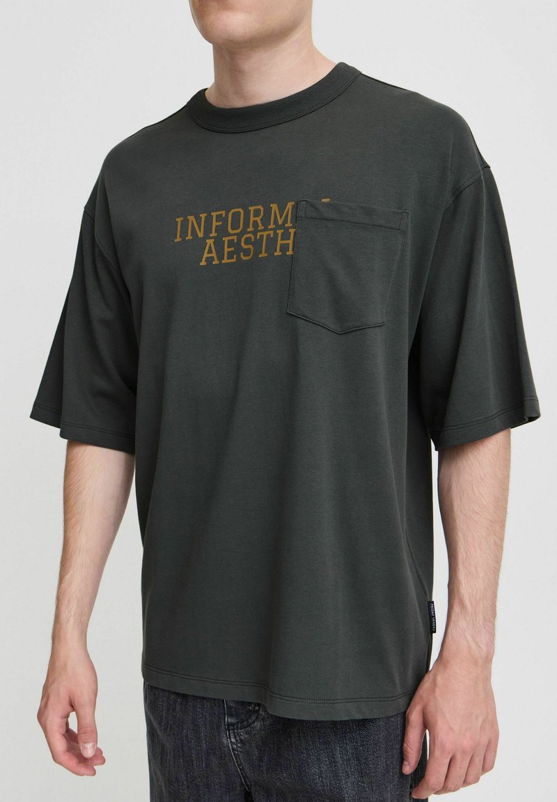 Tamnozelena oversized majica od pamuka s kratkim rukavima, s dijagonalnom chest pocket i natpisom "INFORMAL AESTHETIC" u zlatnoj boji.