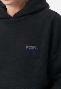 Sweatshirt à capuche en polaire noire, avec un tissu texturé, une poche kangourou à l'avant et un petit logo brodé violet sur la poitrine.