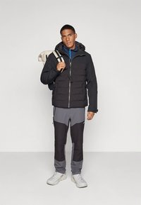 Black insulated jacket with quilted texture, gray pants with color blocking, and white shoes. The model carries a beige backpack with straps.