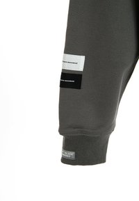 Manga de sudadera gris con puño de canalé. Presenta tres etiquetas de tela en blanco, negro y gris con texto impreso. Textura suave.