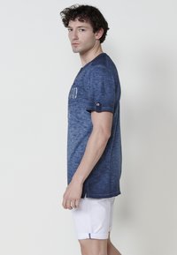 Tummansininen puuvillapaita, jossa on teksturoitu pinta, lyhyet hihat napin koristeilla ja pyöreä pääntie. Yhdistetty valkoisiin shortseihin.