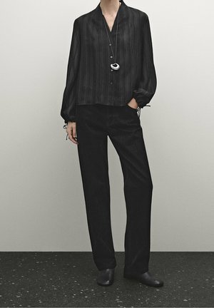 Osoba ubrana w luźną czarną koszulę na guziki, czarne spodnie o prostych nogawkach, czarne buty oraz srebrny naszyjnik z zawieszką, stojąca z ręką w kieszeni.