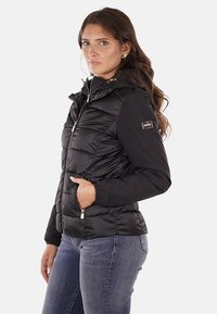 Giacca a vento nera con chiusura a zip, cappuccio e dettaglio di cucitura visibile. Presenta tasche laterali e un'etichetta con logo sulla manica.