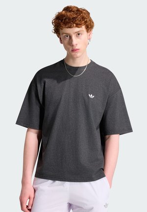 Jeune personne aux cheveux roux bouclés portant un t-shirt Adidas gris foncé et un pantalon blanc, debout devant un fond clair.