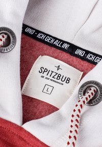 Witte sweatshirt met rode accenten, voorzien van een label met de tekst "SPITZBUB" in het zwart, en een trekkoord met een geweven textuur en metalen oogje.