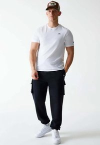 Witte t-shirt met korte mouwen, zwarte cargobroek met zijzakken en witte sneakers. De pet is bruin met een logo. Eenvoudig, casual ontwerp.