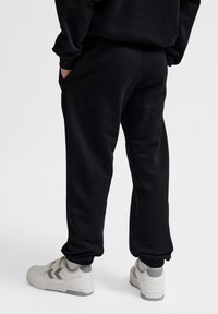 Schwarze Sweatpants mit elastischen Bündchen, seitlichen Taschen und einer lässigen Passform, kombiniert mit weißen und grauen Sneakers. Weicher Stoff.