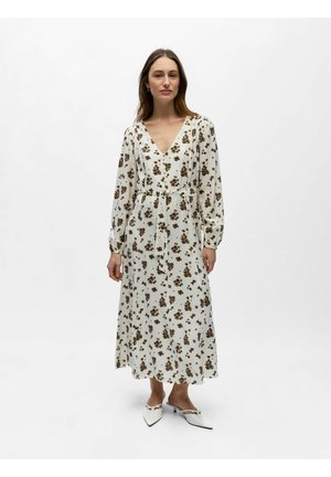 Femme debout portant une robe midi blanche à fleurs, manches longues, col en V, ceinture à la taille, et des escarpins blancs à talons bas et bouts pointus.