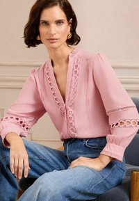 Blusa rosa con dettagli a occhiello lungo il collo e le maniche, caratterizzata da una scollatura profonda a V e polsini arricciati, abbinata a jeans blu.