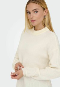 Cremfarbener, gerippter Strickpullover mit hohem Kragen. Weiche Textur mit überschnittenen Schultern und eng anliegenden Bündchen. Auffällig durch schlichtes, elegantes Design.