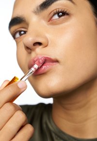 Läppglans i en genomskinlig tube med ett guldlock, applicerat med en flockad applikator. Läpparna är glansiga med en mjuk rosa nyans. Huden är len och strålande.