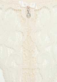 Lingerie en dentelle blanche avec des motifs floraux élaborés, un élément décoratif central avec un pendentif en perle et cristal, et un petit nœud en satin.