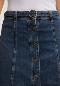 Gonna di jeans con bottoni frontali, realizzata in tessuto blu scuro, con cuciture a contrasto, cintura con fibbia metallica circolare e elementi decorativi in metallo.