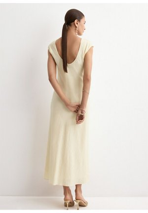 Kobieta z długimi brązowymi włosami związanymi nisko w kucyk, ubraną w kremową, bezrękawową sukienkę midi z głębokim dekoltem na plecach, trzymająca nadgarstki za plecami.