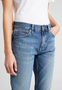 Mujer con jeans de denim de tiro alto de color azul claro y una camiseta blanca metida por dentro, con una mano en el bolsillo y la otra relajada a su lado.