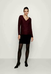 Top lung cu mânecă lungă de culoare burgundă, cu decolteu în V, purtat cu o fustă mini neagră și colanți negri. Modelul poartă pantofi stil stiletto negri.