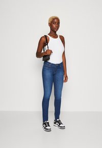 Lieknos mėlynos džinsinio audinio kelnės su vidutinio aukščio juosmeniu, derinamos su baltu raštuotu tank topu ir juodais sportbačiais. Modelis laiko mažą juodą rankinę.