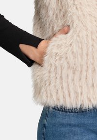 Gilet en fausse fourrure beige clair, avec une texture douce et duveteuse. Associé à une manche noire présentant un détail découpé au niveau du poignet.