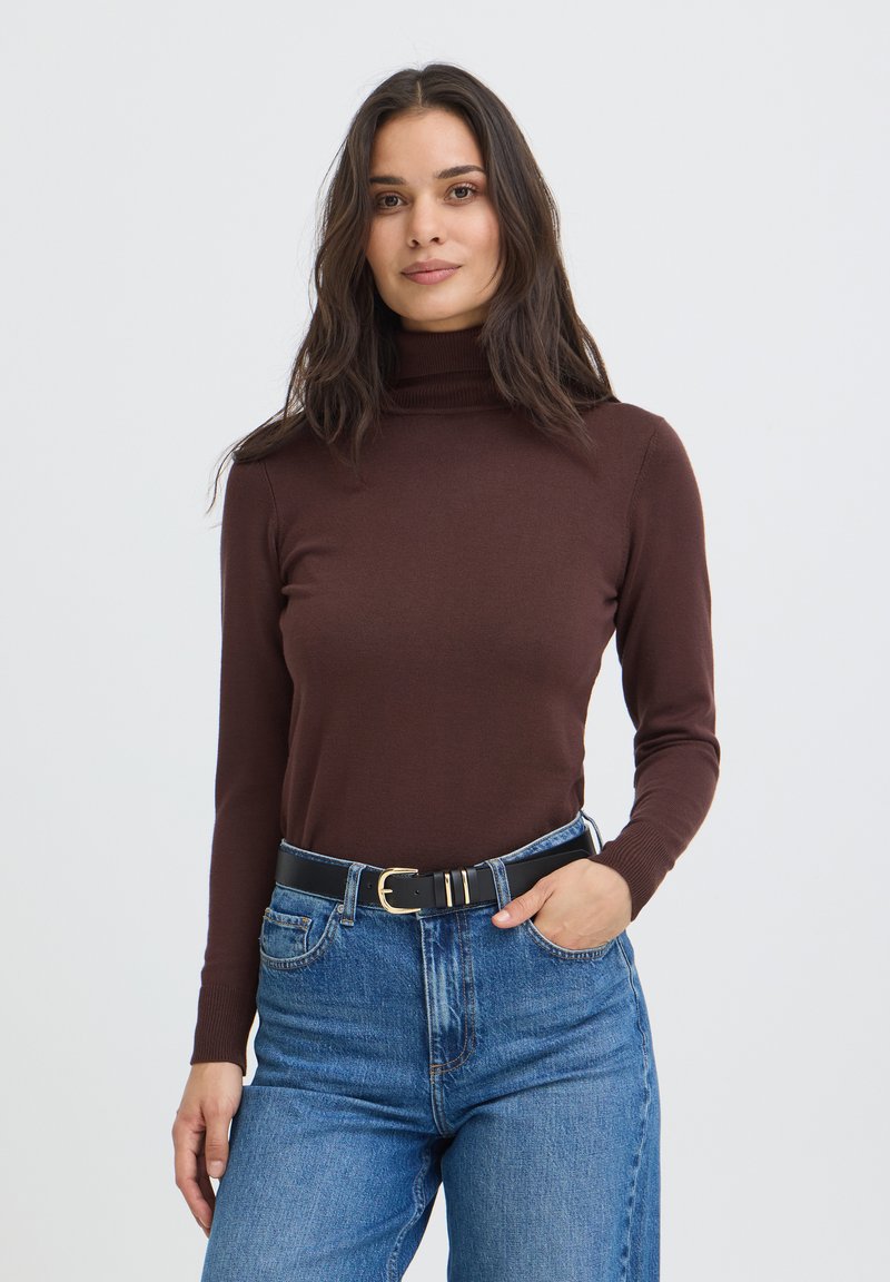 Femme aux longs cheveux foncés portant un pull à col roulé marron, un jean bleu et une ceinture noire, debout avec une main dans la poche.