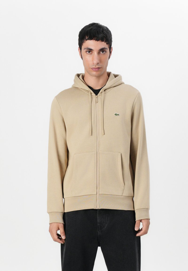 Felpa beige con zip, realizzata in un tessuto morbido. Presenta un cappuccio, tasche frontali e un logo verde ricamato sul lato sinistro del petto.