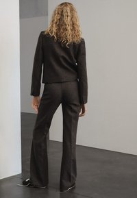 Completo in lana marrone con giacca su misura e pantaloni a campana. Il tessuto ha una finitura textured. L'outfit è abbinato a scarpe scure.