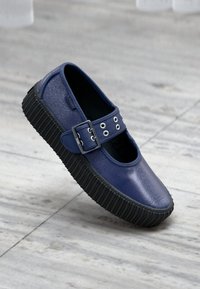 Donkerblauwe Mary Jane-stijl schoen met een textuur bovenwerk, gesp-detail en dikke rubberen zool, gepresenteerd tegen een lichte vloer.