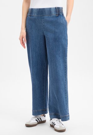 Niebieskie dżinsy o szerokich nogawkach z elastycznym pasem, noszone z białymi i czarnymi sneakersami, pokazane od talii do stóp.