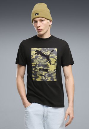 Ung mand iført en sort Puma t-shirt med gul camouflageprint og en gul hue, stående med den ene hånd i lommen mod en grå baggrund.