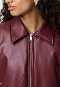 Burgundy faux läderjacka med en stor krage, silverdragkedja och slät textur, som visar en del av en persons ansikte med lockigt hår.