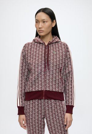 Vrouw draagt een bordeauxrode capuchontrui met rits en een bijpassende broek met gestreepte mouwen en broekzijden, staand tegen een effen achtergrond.