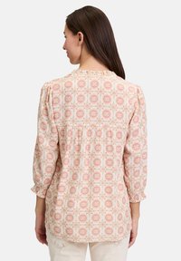 Blusa a fantasia con maniche a tre quarti, caratterizzata da un collo a volant, di colore rosa pallido con motivi floreali in tonalità arancione, gialla e blu.