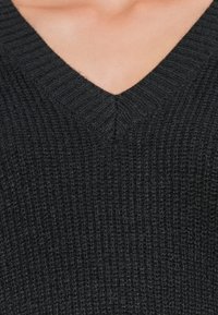 Gros plan sur le haut de la poitrine portant un pull en maille noire à col en V avec un motif côtelé texturé.