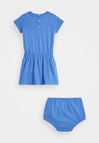 Blå bomullsklänning med korta ärmar, elastisk midja och knäppning bak. Matchande blå bomullsshorts ingår. Slät yta och avslappnad design.