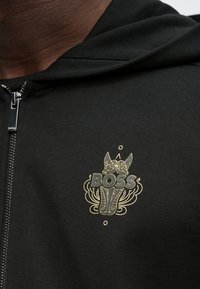Camisola com fecho de correr preta, com um remendo gráfico de um cão e a palavra "BOSS" em letras douradas, ousadas e ornamentadas. Textura suave.