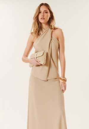Femme en top halter beige et longue jupe tenant une pochette assortie, accessoirisée de bracelets épais, sur fond blanc uni.