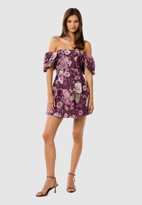 Vrouw in een paarse off-shoulder mini-jurk met roze en beige bloemenpatroon, gecombineerd met bruine gestructureerde hoge hakken.