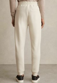 Pantaloni de trening bej deschis, cu talie elastică, buzunare laterale și glezne mansetate. Textură netedă, asortați cu încălțăminte închisă la culoare.