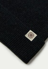 Svart stickad mössa med ribbat mönster och en liten krämfärgad etikett med en brun dekorativ logotyp broderad på kanten.