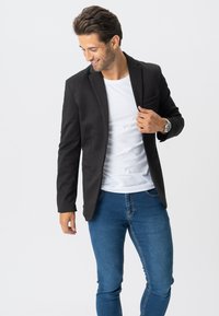 Sort blazer lavet af vævet stof, slim fit, med enkeltknaplukning, to frontlommer, båret over en hvid t-shirt og blå jeans.