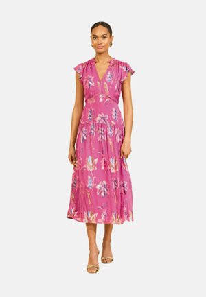 Femme portant une robe midi rose à imprimé floral, manches volantes, décolleté en V et talons ouverts dorés, debout sur fond blanc.