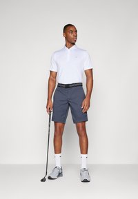 Hvit polo skjorte med krage, grå shorts, svart belte, grå joggesko og hvite sokker. Holder en golfklubbe og står avslappet.