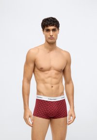 En mann uten skjorte med kort krøllete hår, iført røde mønstrede Calvin Klein boxershorts, mot en enkel lys bakgrunn.