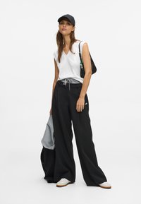 Λευκή αμάνικη μπλούζα με v-λαιμόκοψη, φαρδιά μαύρα παντελόνια με κορδόνι, γκρι πουλόβερ ριγμένο, μαύρο καπέλο, μαύρη τσάντα και λευκά αθλητικά παπούτσια με καφέ σόλες.