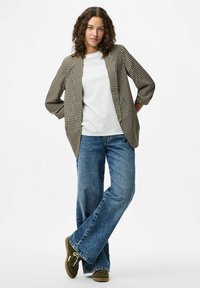 Mlada žena s kovrčavom kosom obučena u blejzer s uzorkom hounds-tooth, bijelu košulju, široke plave traperice i zelene tenisice, stoji s prekrštenim nogama.