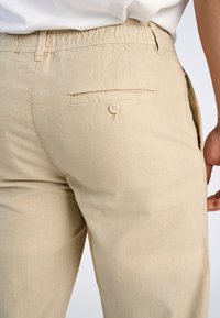 Beige linnen broeken met een knoopsluiting achtervak, elastische tailleband en een gladde textuur, zij-aan-zij getoond.