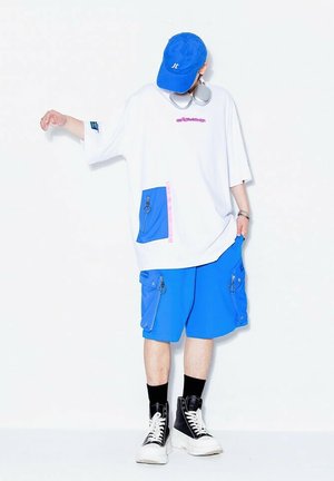 Camiseta blanca de gran tamaño con detalles en rosa y azul, combinada con pantalones cortos azules con bolsillos con cremallera, y zapatillas de caña alta negras.