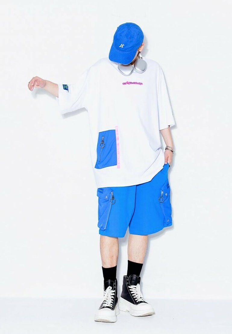 Camiseta blanca de gran tamaño con detalles en rosa y azul, combinada con pantalones cortos azules con bolsillos con cremallera, y zapatillas de caña alta negras.